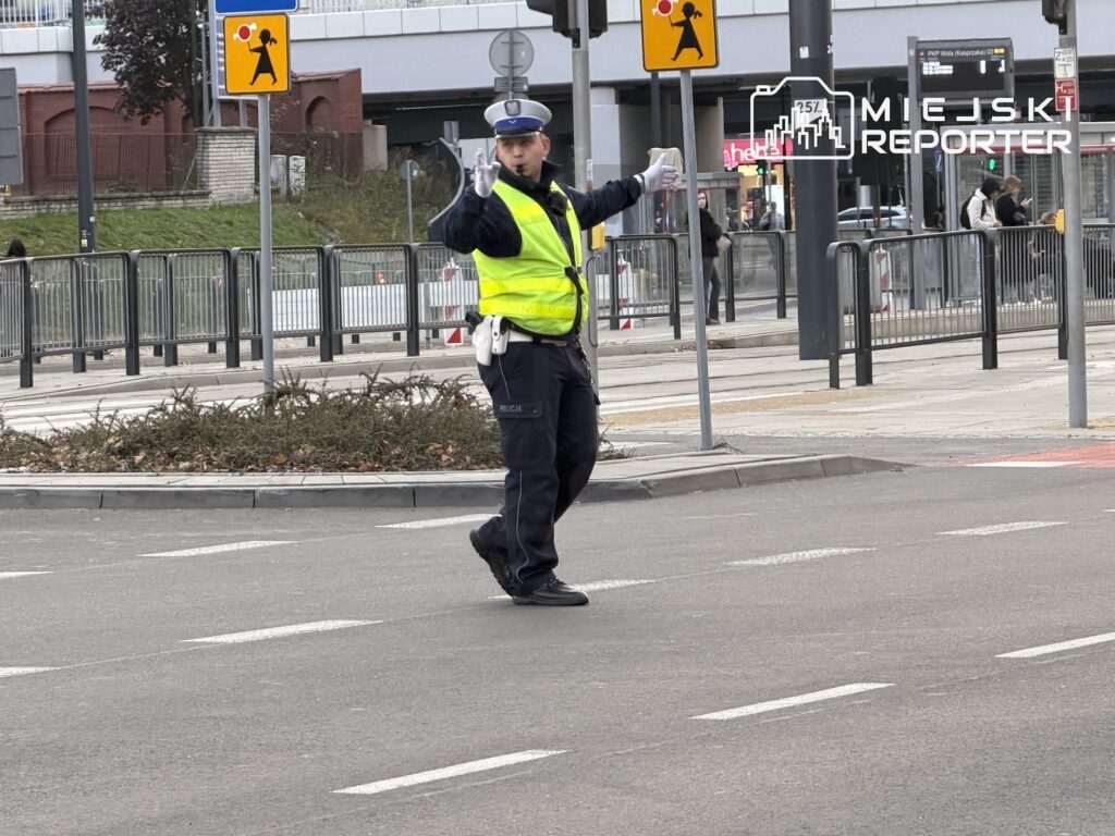 Policjant w odblaskowej kamizelce kieruje ruchem na skrzyżowaniu z torowiskiem tramwajowym.
