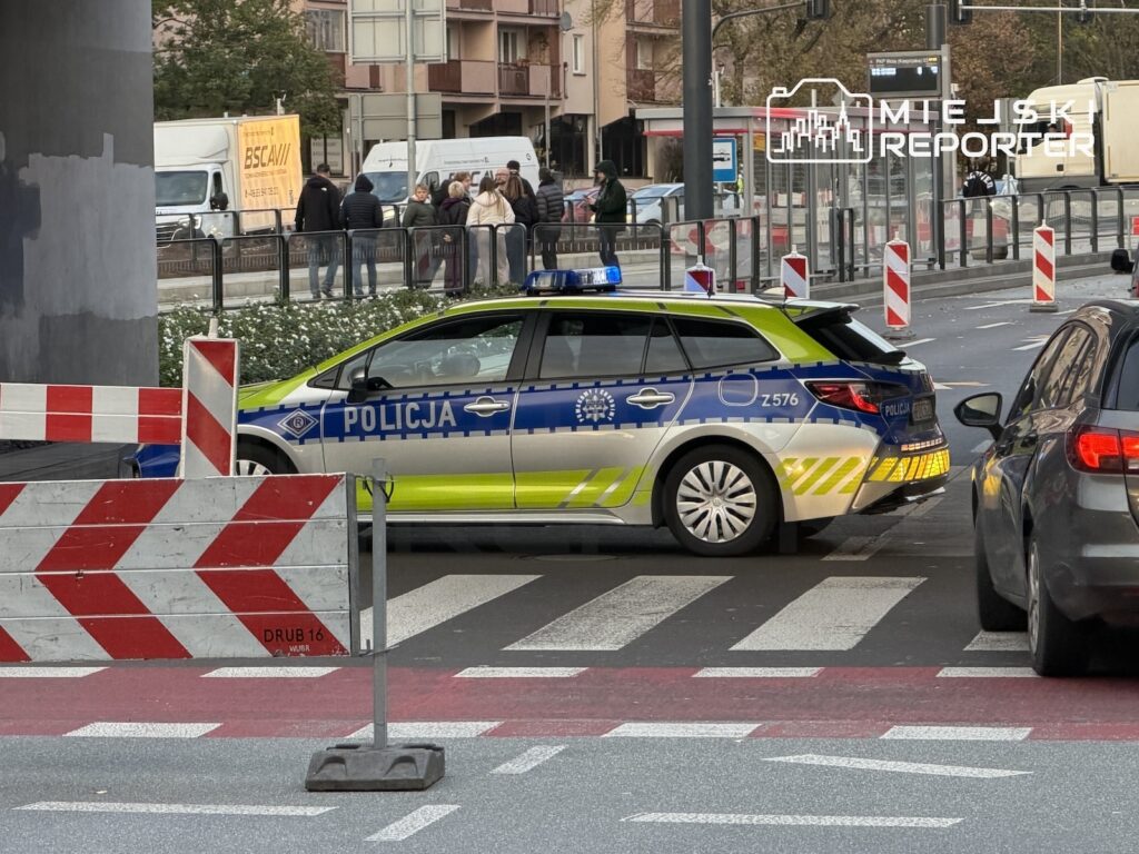 Oznakowany radiowóz policyjny stoi na przejściu dla pieszych przy zwężonym odcinku jezdni z barierami drogowymi.