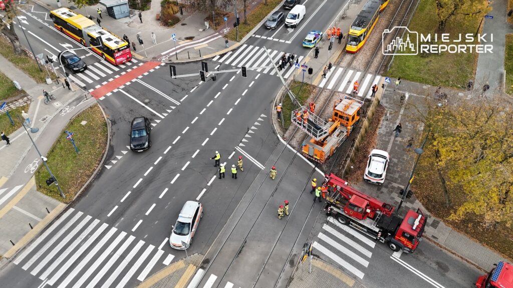Strażacy i służby drogowe pracują przy torowisku na skrzyżowaniu ulic, obok wozu strażackiego i tramwaju technicznego.