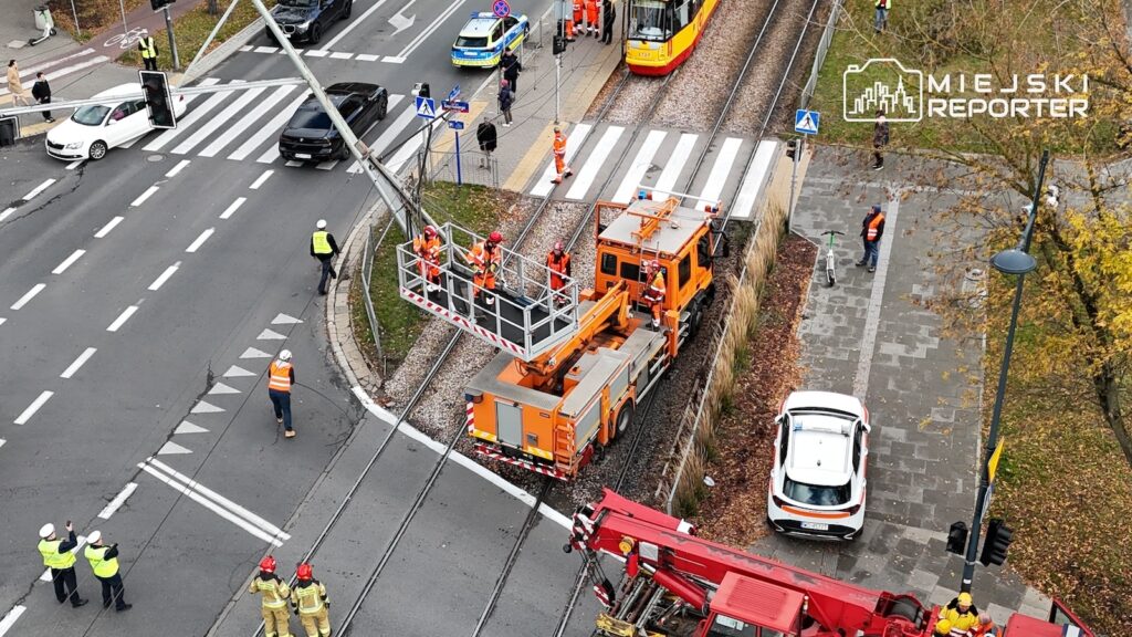 Ekipy techniczne i strażacy pracują przy pomarańczowym podnośniku na torowisku, obok skrzyżowania i tramwaju.