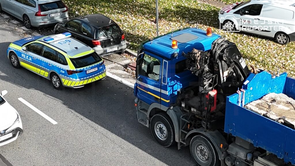 Radiowóz policji stoi obok niebieskiej ciężarówki z podniesionym mechanizmem, na parkingu przy zaparkowanych samochodach.