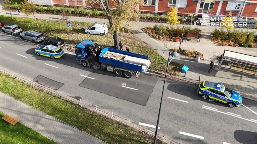 Dwa oznakowane radiowozy policyjne i niebieska ciężarówka stoją na ulicy przed blokiem mieszkalnym.