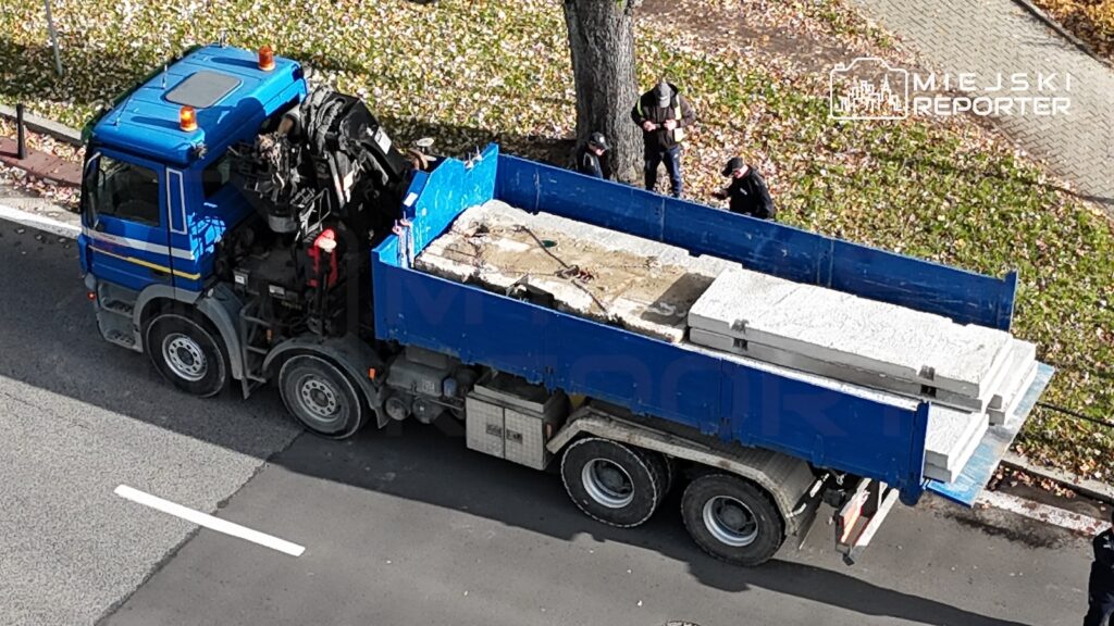 Niebieska ciężarówka z dźwigiem załadowana betonowymi elementami stoi na ulicy przy chodniku.
