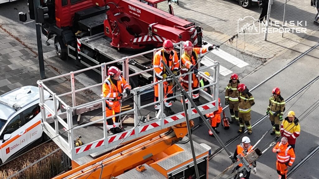 Strażacy i pracownicy w odblaskowych kombinezonach pracują przy podnośniku koszowym na ulicy.