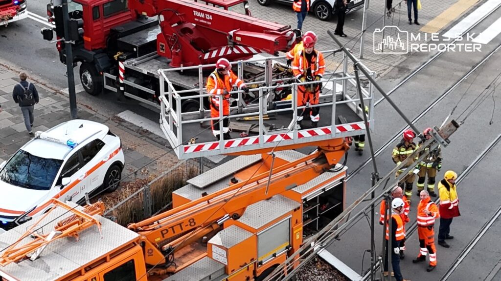 Strażacy pracują na podnośniku przy sieci trakcyjnej nad torowiskiem, obok stoją wozy ratownicze.