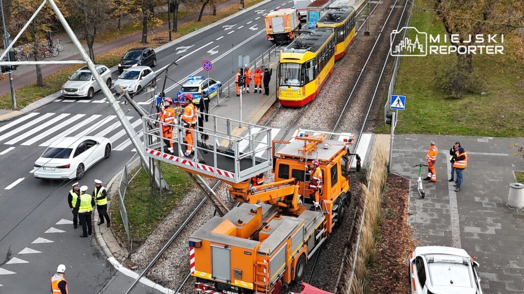 Pomarańczowy pojazd techniczny z podnośnikiem pracuje przy torowisku obok żółto-czerwonego tramwaju i ekipy w kamizelkach