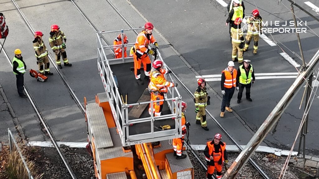 Strażacy i pracownicy służb technicznych pracują przy podnośniku na torowisku, obok którego stoją wozy i sprzęt ratowniczy.