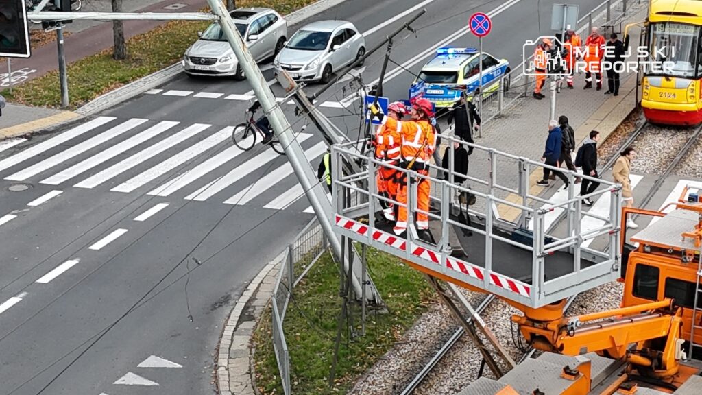Pracownicy w pomarańczowych uniformach na podnośniku naprawiają sieć trakcyjną nad torowiskiem tramwajowym.