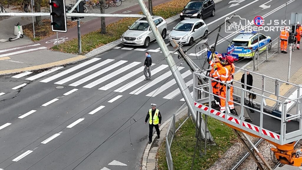 Pracownicy w pomarańczowych kombinezonach pracują na podnośniku przy skrzyżowaniu, gdzie ruch zabezpiecza policjant.