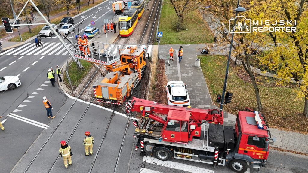 Strażacy i ekipy techniczne pracują przy czerwonym wozie z podnośnikiem obok tramwaju na torowisku.