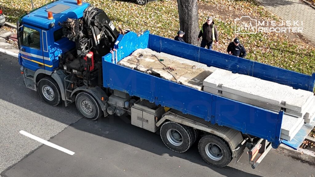 Niebieska ciężarówka z dźwigiem przewozi betonowe elementy, obok stoją dwie osoby.