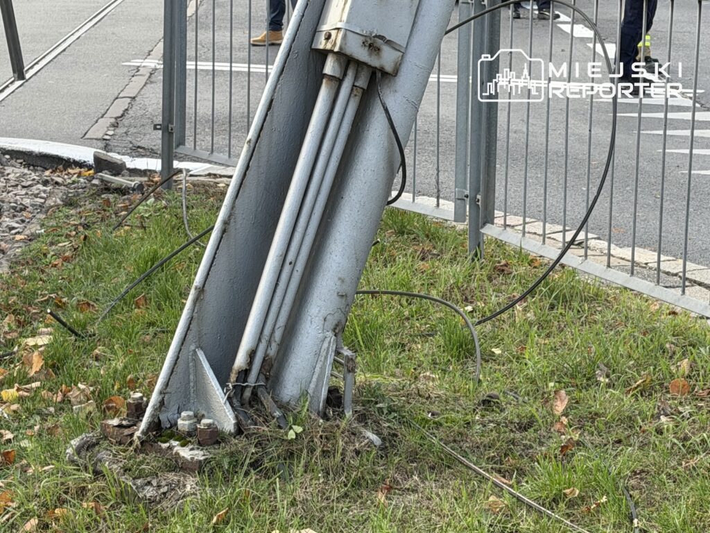 Pochylony stalowy słup z przewodami przy ogrodzeniu, na trawiastym skraju ulicy.