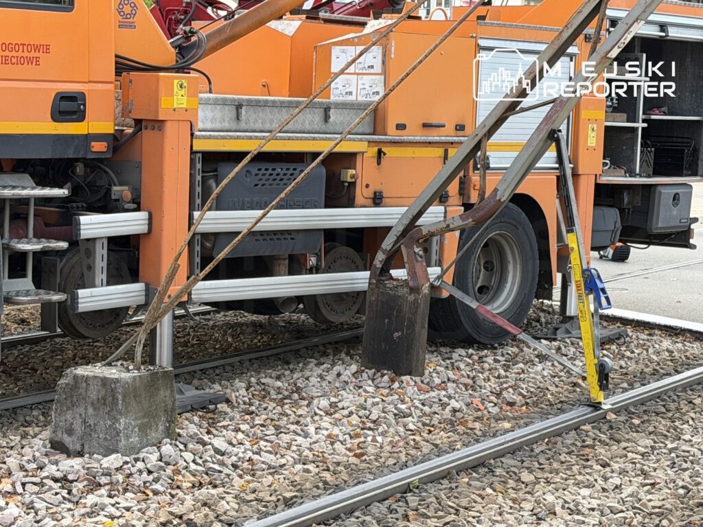 Pomarańczowy pojazd techniczny stoi na torach kolejowych przy metalowych podporach i sprzęcie zabezpieczającym.