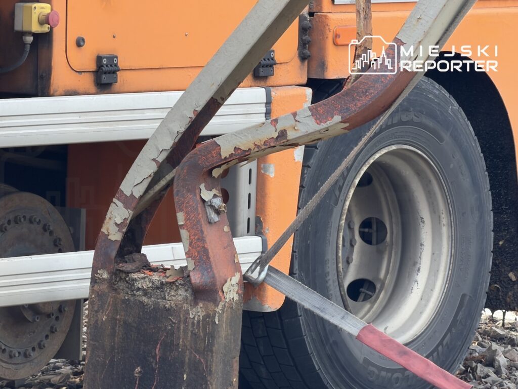 Zardzewiały metalowy element leży na gruzie przy kole pomarańczowego pojazdu ciężarowego.