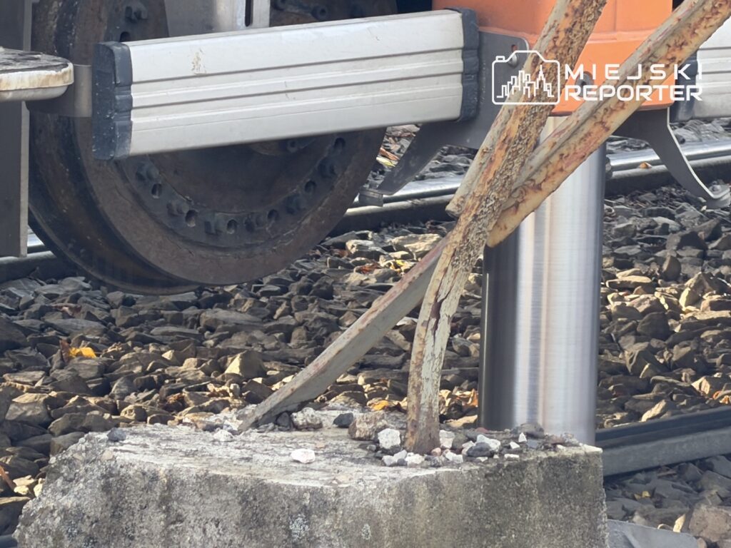 Koło lokomotywy nad torowiskiem, obok stalowego słupka i zardzewiałego wspornika osadzonego w betonowej podstawie.