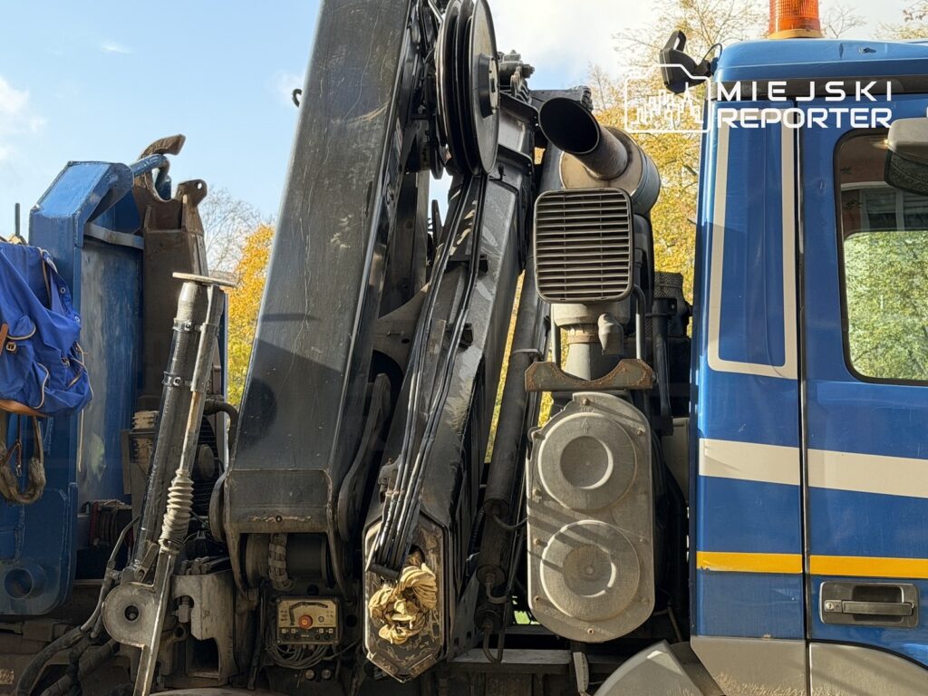 Zbliżenie na mechaniczne ramię i osprzęt niebieskiej koparki zaparkowanej pod drzewami.