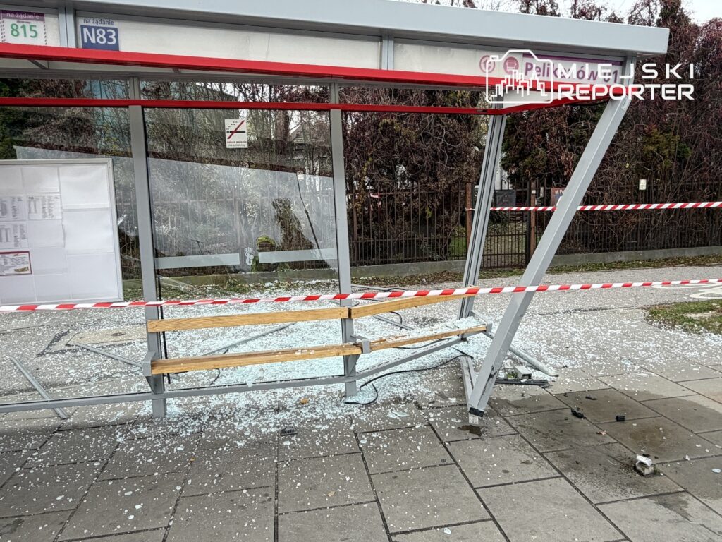 Uszkodzona wiata przystankowa z rozbitą szybą i odłamkami szkła na chodniku, zabezpieczona biało-czerwoną taśmą.