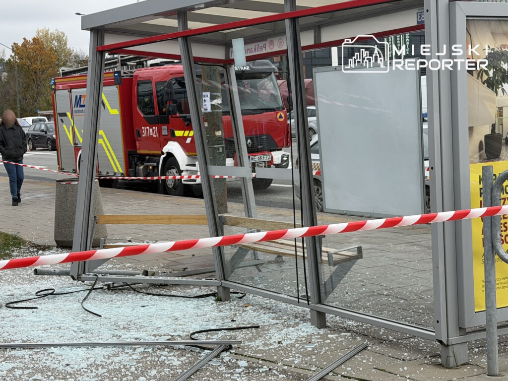 Rozbita szklana wiata przystankowa odgrodzona taśmą, za nią stoją wozy strażackie.