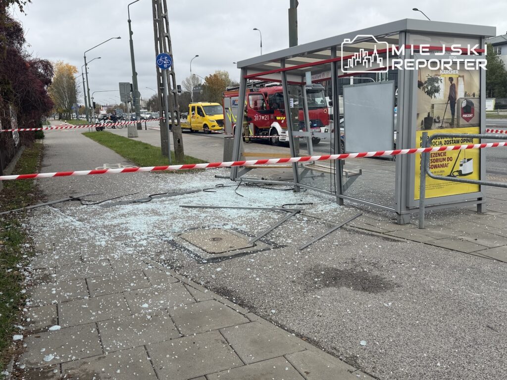Uszkodzona wiata przystankowa otoczona taśmą, z rozbitym szkłem na chodniku i wozem strażackim w tle.