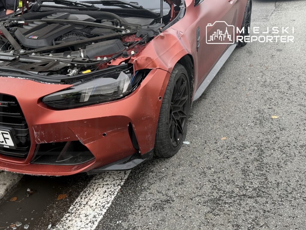 Czerwony samochód z otwartą maską i uszkodzonym przednim błotnikiem stoi na jezdni przy białej linii.