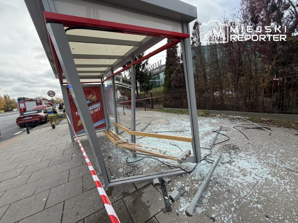 Uszkodzona wiata przystankowa z rozbitymi szybami, otoczona taśmą, przy chodniku obok stojących pojazdów.