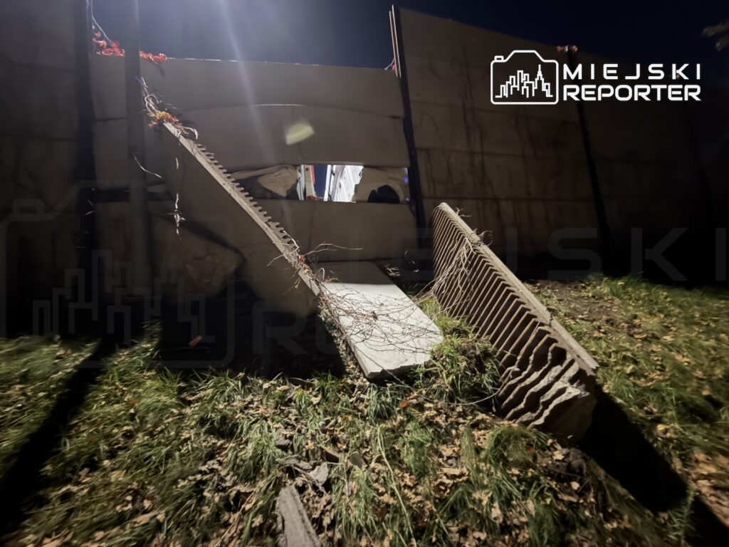 Połamane betonowe elementy leżą na trawie przy uszkodzonym murze, nocą oświetlone reflektorem.