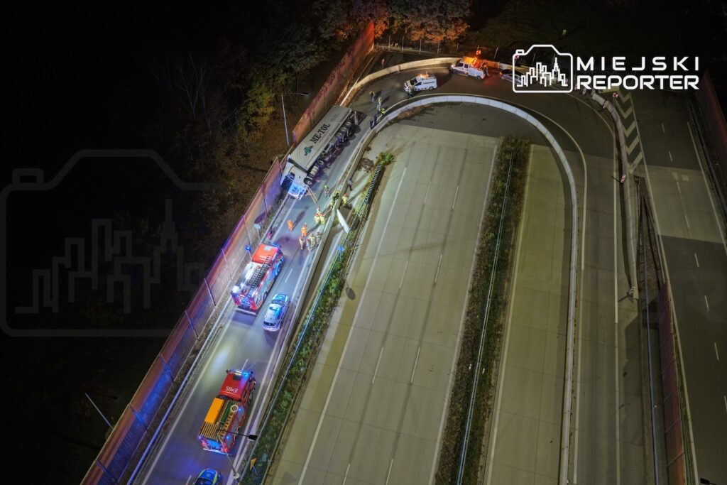 Widok z drona na nocną akcję służb przy drodze; pojazdy z włączonymi sygnałami świetlnymi stoją wzdłuż jezdni.