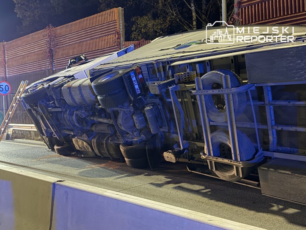 Przewrócona ciężarówka leży na boku na jezdni przy barierach, nocą oświetlona niebieskim światłem.