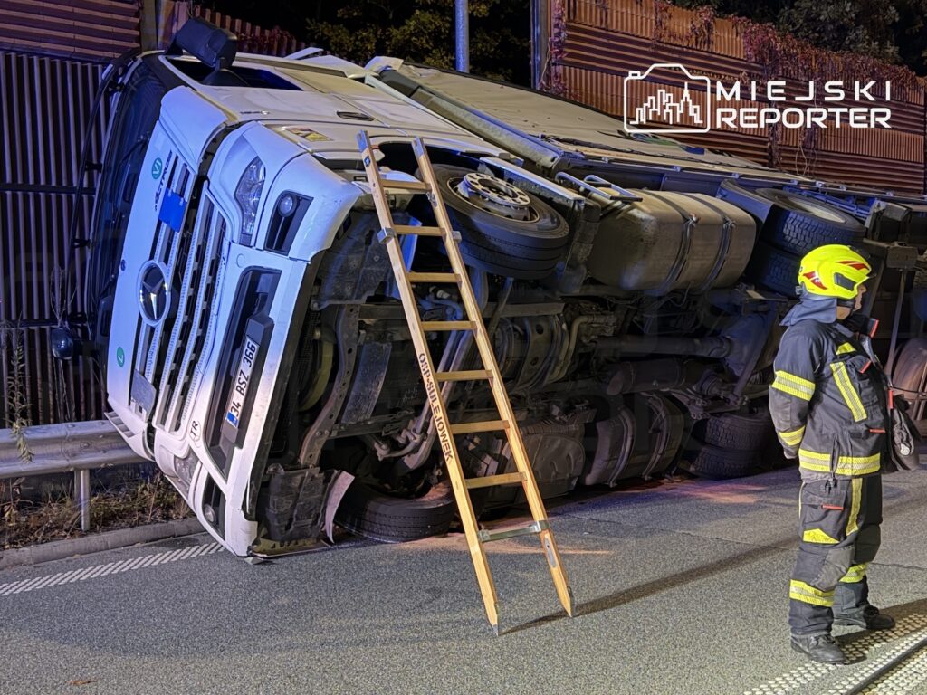 Ciężarówka leży na boku przy ogrodzeniu, a strażak w kasku ogląda pojazd obok rozstawionej drabiny.