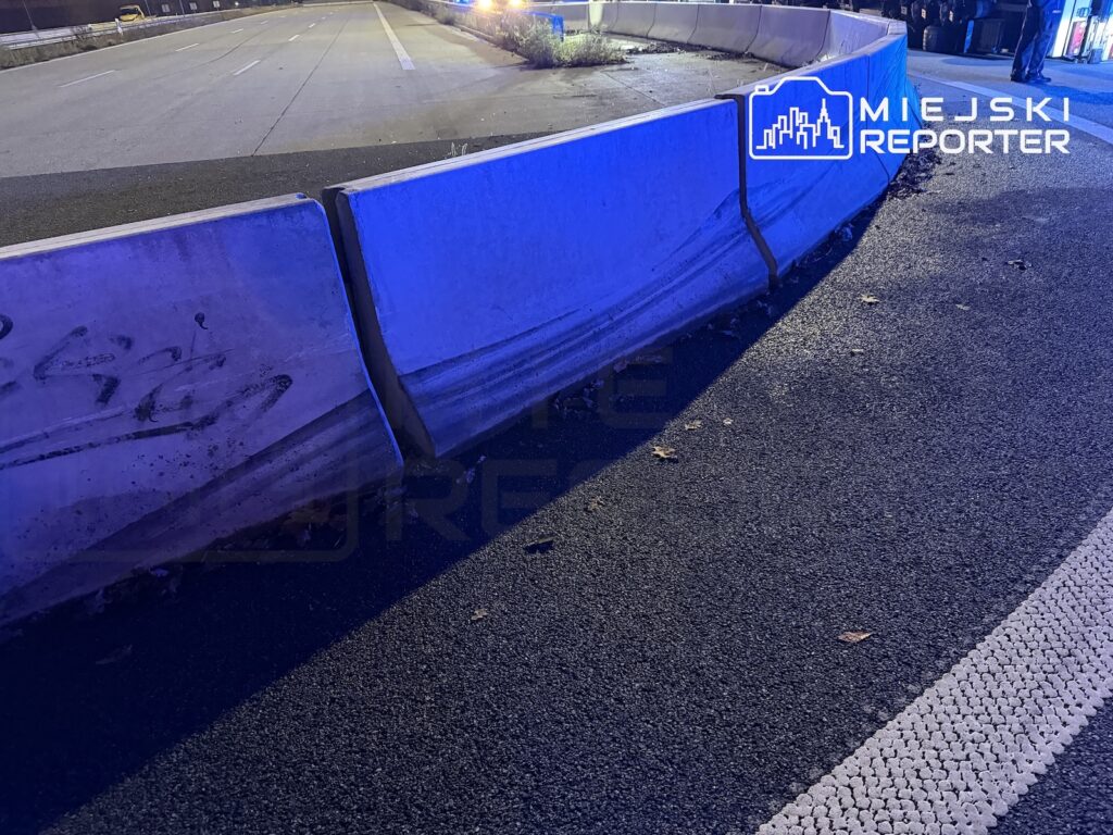 Betonowe bariery drogowe zamykają fragment jezdni nocą, oświetlone niebieskim światłem.