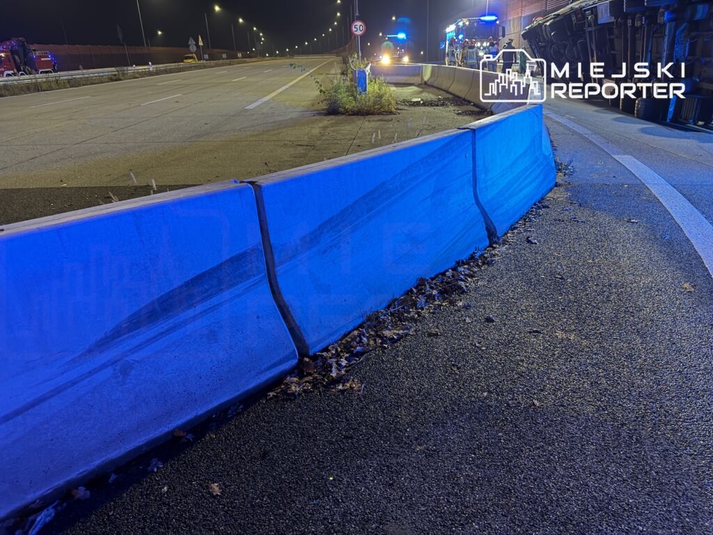 Niebieskie bariery oddzielają jezdnię od chodnika na nocnej drodze, obok stoją pojazdy służb z włączonymi sygnałami