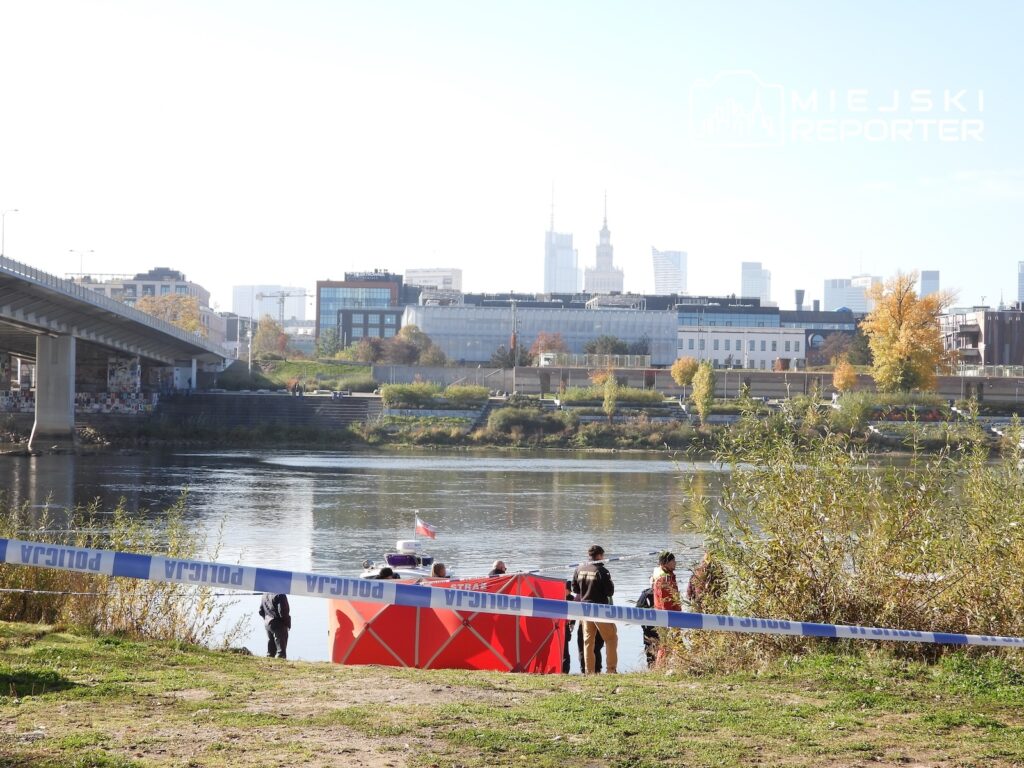 Służby przy czerwonej łodzi nad brzegiem rzeki, za taśmą zabezpieczającą, na tle mostu i miejskiej zabudowy.