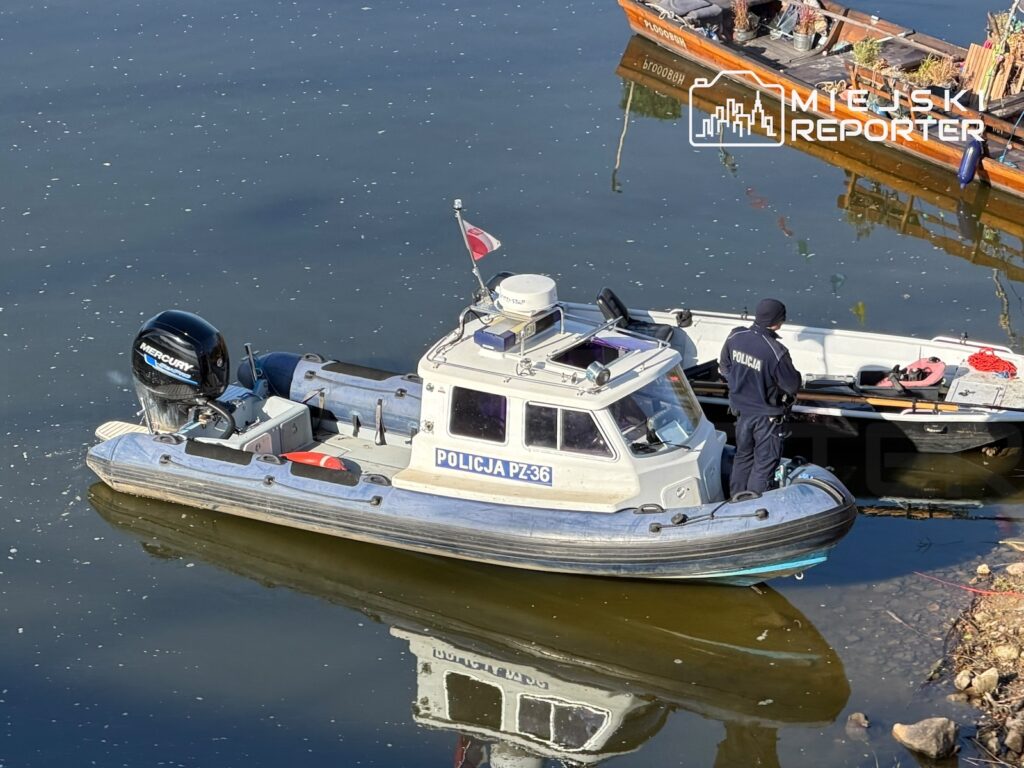 Policyjna łódź z funkcjonariuszem na pokładzie stoi na rzece przy brzegu, obok drugiej łodzi.