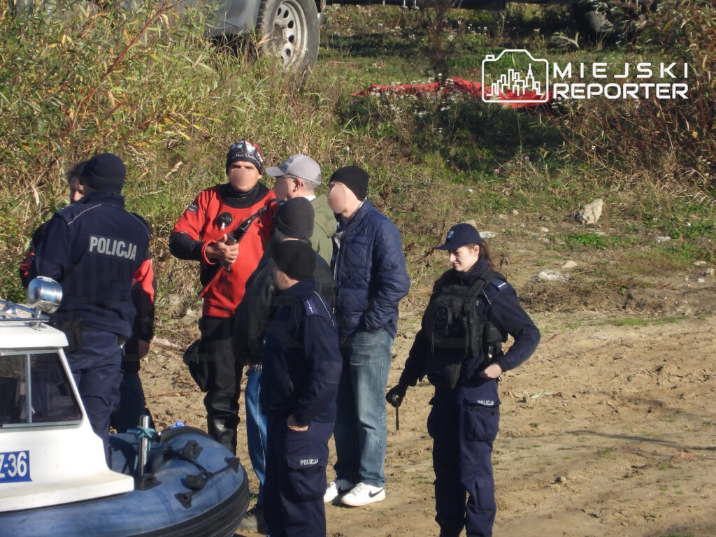 Policjanci i mężczyźni w cywilnych ubraniach rozmawiają na brzegu rzeki obok łodzi służbowej.