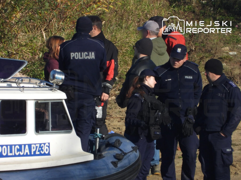 Policjanci i cywile stoją przy łodzi policyjnej na brzegu rzeki, wśród zarośli.