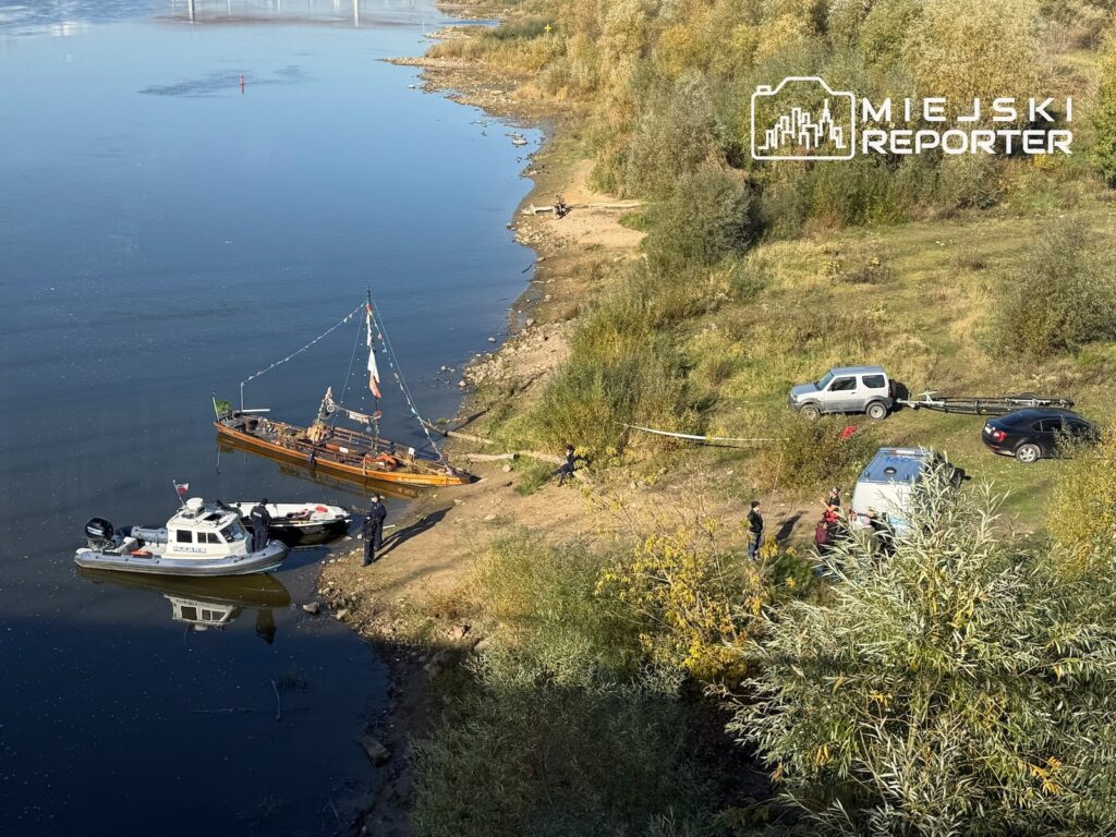 Łodzie i służby przy brzegu rzeki, obok których na zarośniętym terenie stoją samochody.