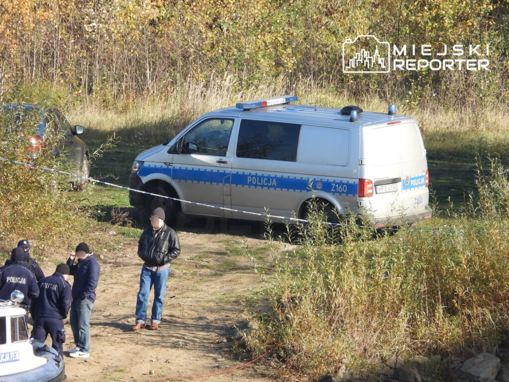 Policyjny bus zaparkowany na zarośniętym brzegu rzeki, obok policjanci i taśma zabezpieczająca teren.