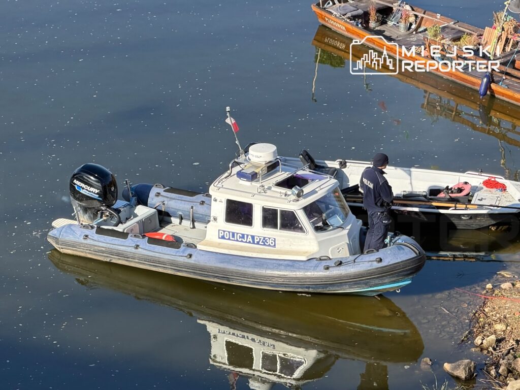 Policyjna łódź motorowa z funkcjonariuszem na pokładzie przy brzegu rzeki, obok zacumowanych łodzi.