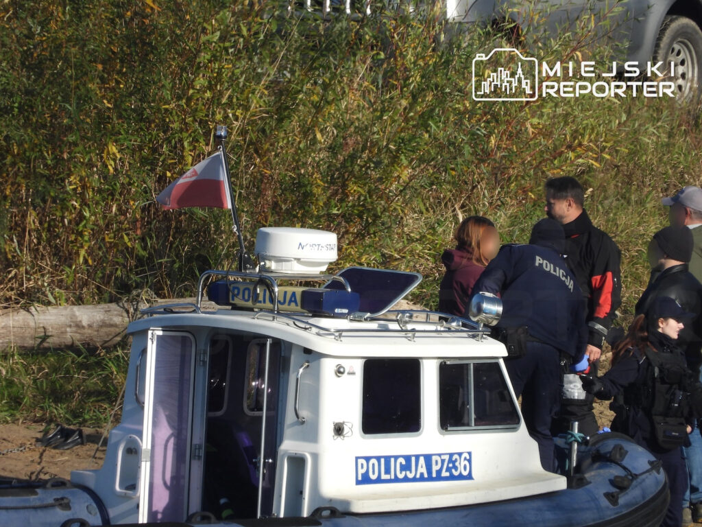 Policjanci stoją przy łodzi policyjnej oznaczonej „POLICJA PZ-36”, na brzegu porośniętym trzcinami.