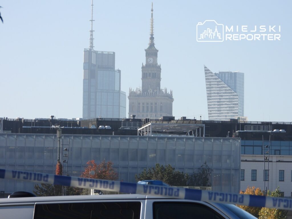 Taśma policyjna na pierwszym planie, a za nią zabudowania i wieżowce na tle Pałacu Kultury i Nauki w Warszawie.