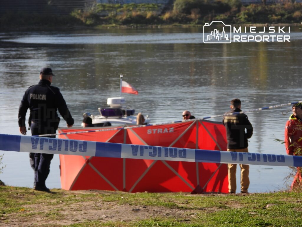 Policjanci i strażacy zabezpieczają brzeg rzeki taśmą policyjną przy czerwonym namiocie służb ratunkowych.