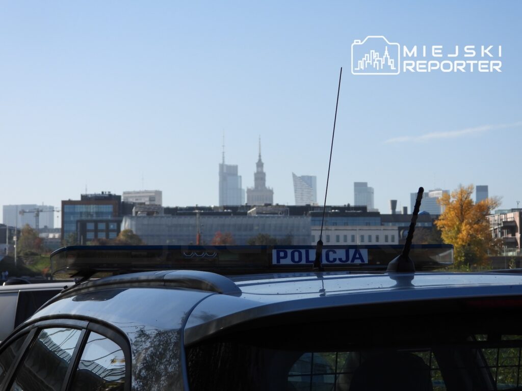 Radiowóz z napisem „POLICJA” na tle warszawskich wieżowców i jesiennych drzew.