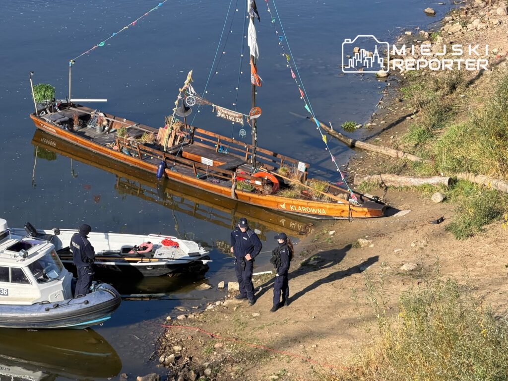 Funkcjonariusze stoją przy łodziach służbowych i drewnianej barce zacumowanej przy brzegu rzeki.