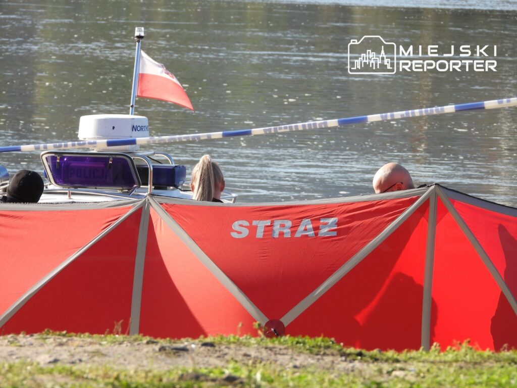 Czerwony parawan z napisem „STRAŻ” osłania służby i łódź przy brzegu rzeki; w tle widać taśmę zabezpieczającą.