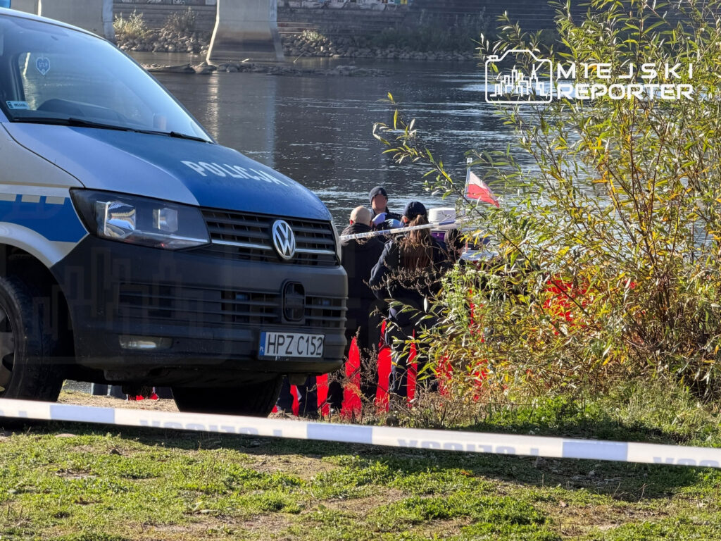 Policyjny van i funkcjonariusze za taśmą zabezpieczającą nad brzegiem rzeki, wśród zarośli.
