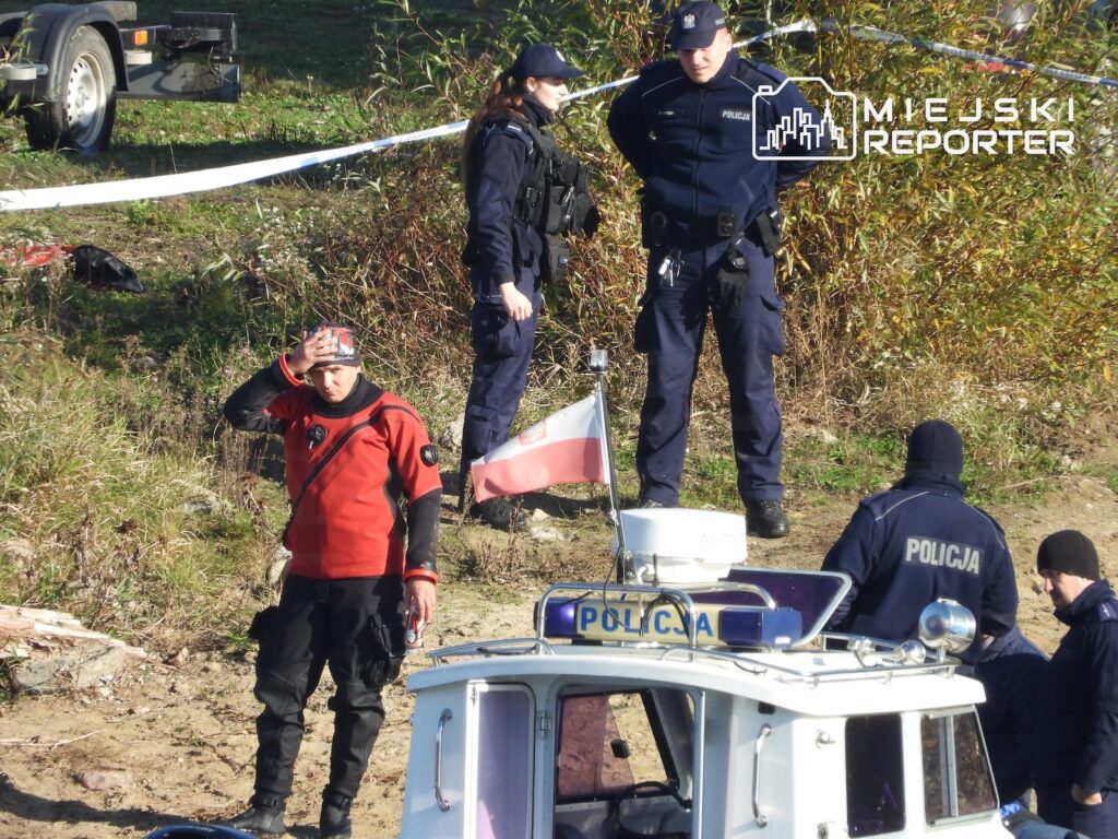 Policjanci i ratownik w czerwonej kurtce stoją na zarośniętym brzegu rzeki obok łodzi policyjnej.