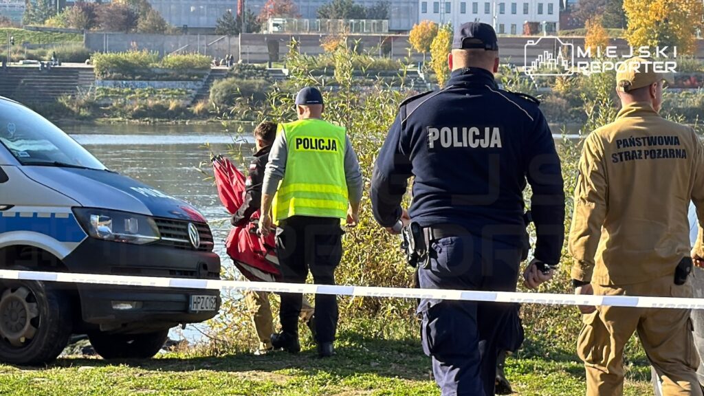 Policjanci i strażak stoją za taśmą zabezpieczającą nad brzegiem rzeki, obok radiowozu.