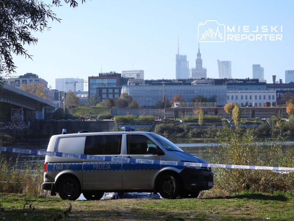 Radiowóz policyjny za taśmą zabezpieczającą nad rzeką, na tle miejskiej zabudowy.