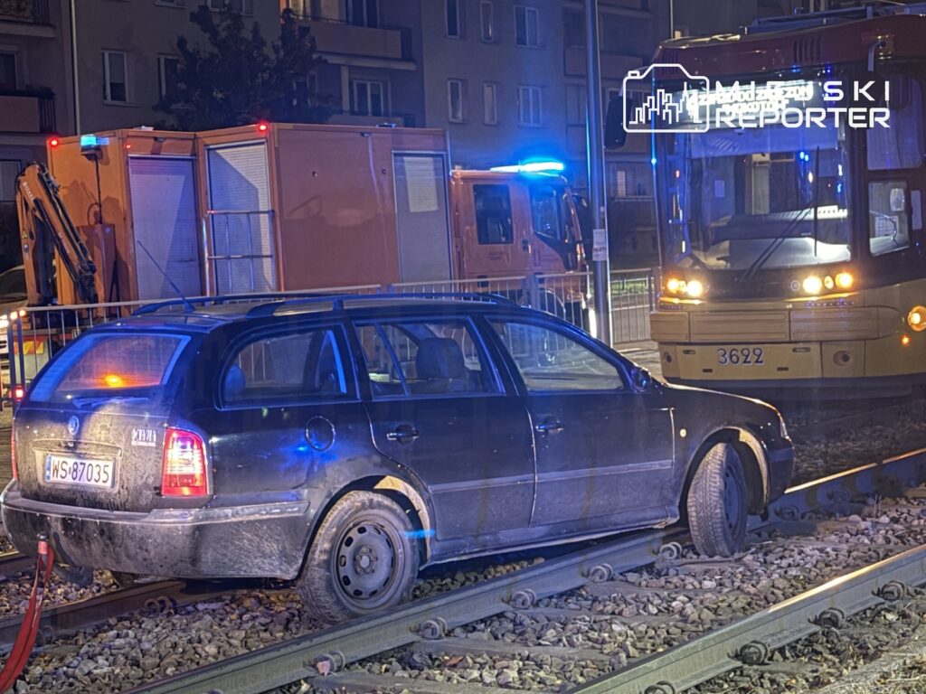 Uszkodzony samochód osobowy stoi na torowisku przed tramwajem i pojazdem służb ratunkowych nocą.