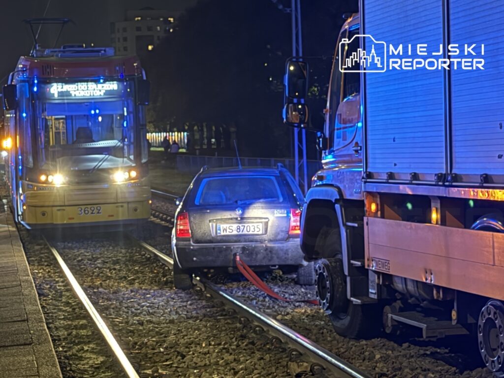 Samochód osobowy stoi na torowisku między tramwajem a ciężarówką, nocą na miejskiej ulicy.